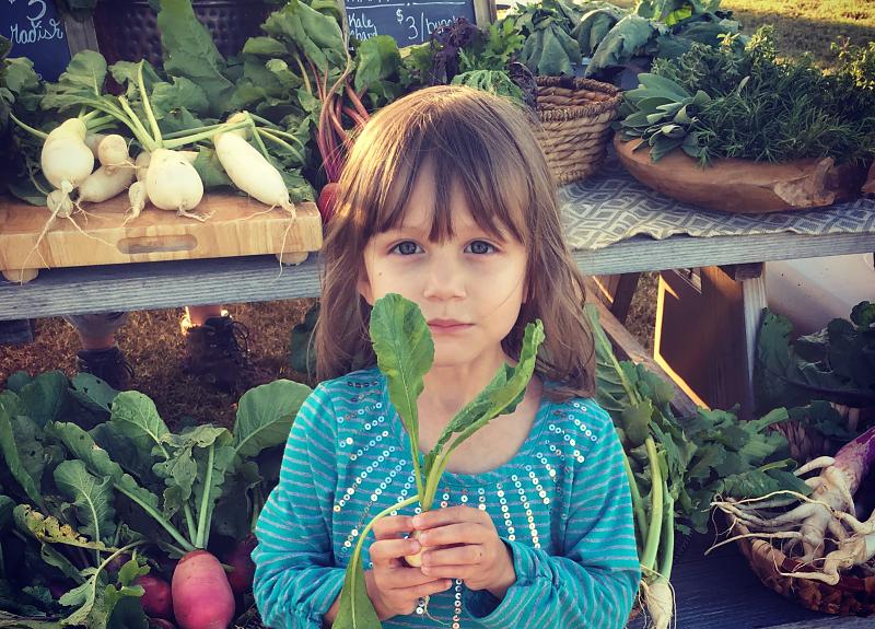 Girl poses with produce at an organic produce stand in Fort Bend.