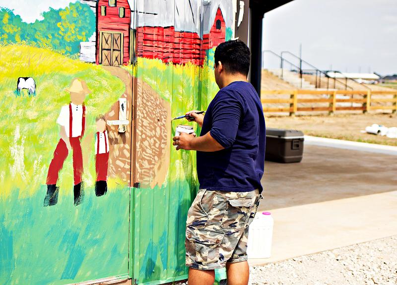 A vibrant, colorful farmhouse scene mural is being painted in Harvest Green.